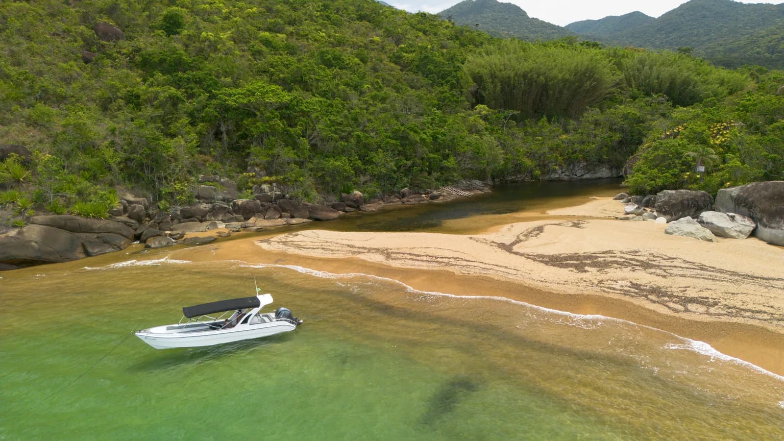Praia cercada por vegetação e lancha sobre água rasa, vista do alto